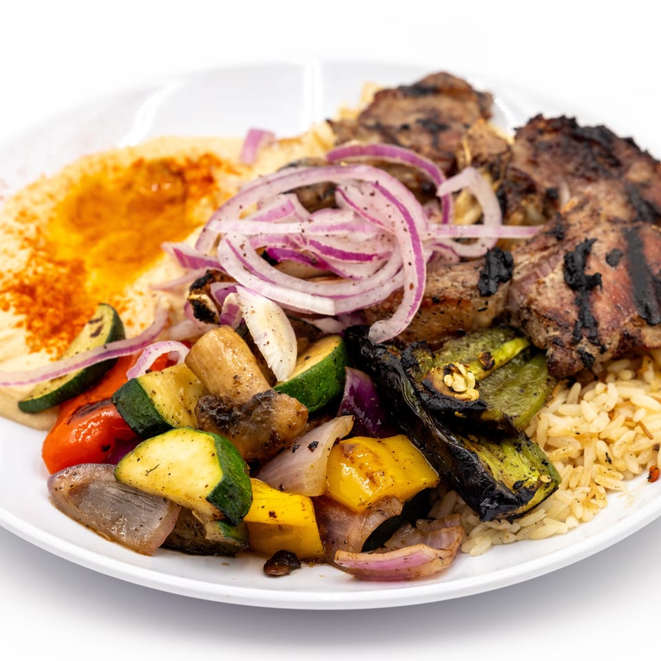 Plate of grilled meat, roasted vegetables, rice, hummus, and sliced red onions.