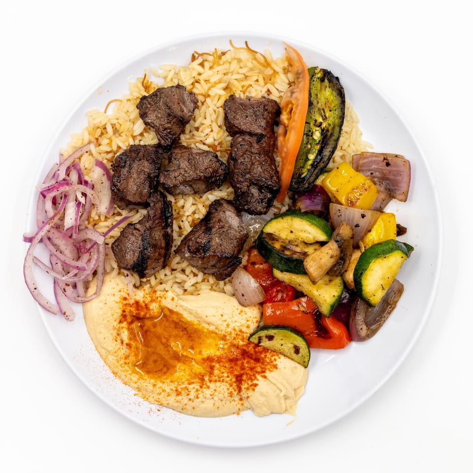 Plate of grilled beef kebab pieces with rice, hummus, grilled vegetables, and sliced red onions.