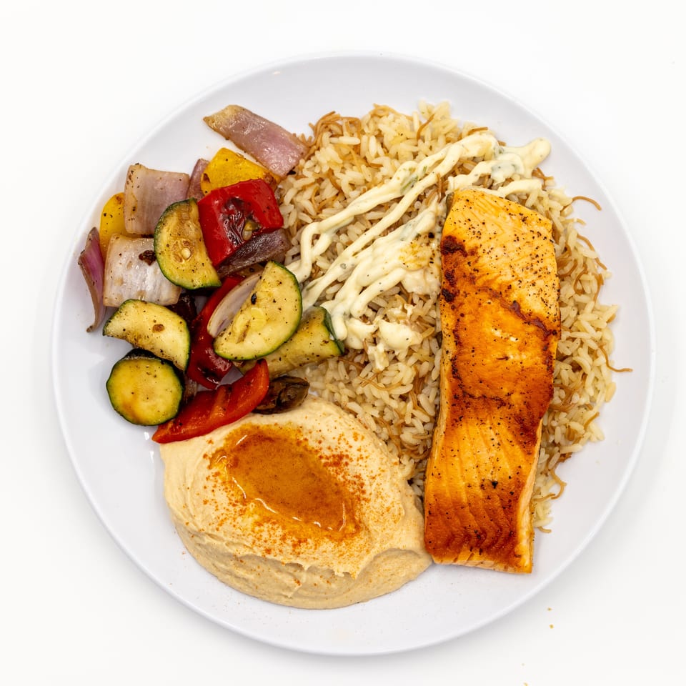 Plate of grilled salmon with rice, hummus, creamy sauce, and roasted zucchini, peppers, onions, and mushrooms.