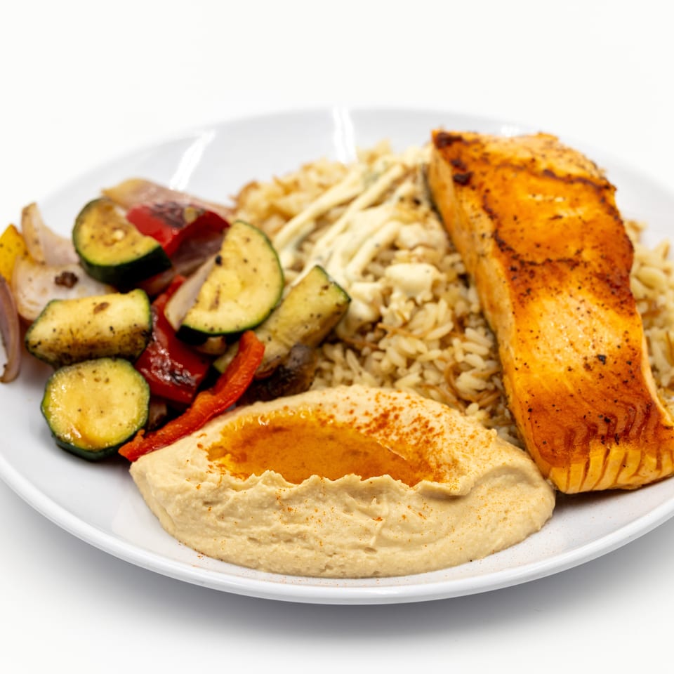 Plated meal featuring grilled salmon, creamy hummus, rice pilaf, and roasted zucchini, peppers, onions, and eggplant.