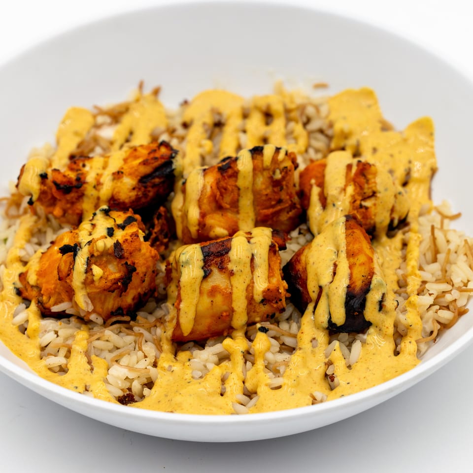 Grilled chicken pieces drizzled with creamy yellow sauce over seasoned rice in a white bowl.