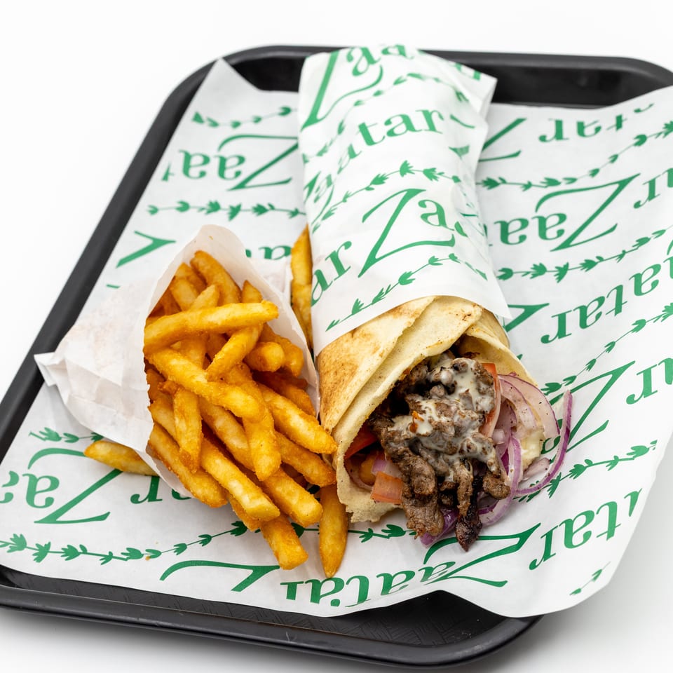 Gyro sandwich with grilled meat, onions, tomatoes, and sauce served alongside golden fries on a branded paper-lined tray.