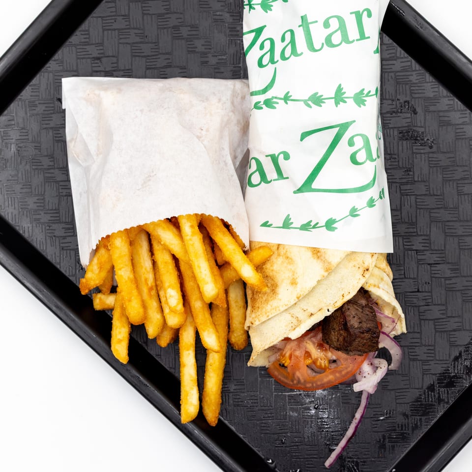 Gyro wrapped in branded paper with grilled meat, tomato, and onion, served alongside golden fries on a black tray.