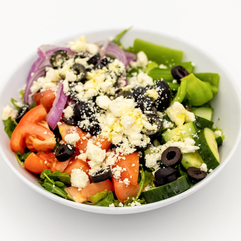 Greek salad with lettuce, tomatoes, cucumbers, red onions, black olives, and crumbled feta cheese in a white bowl.