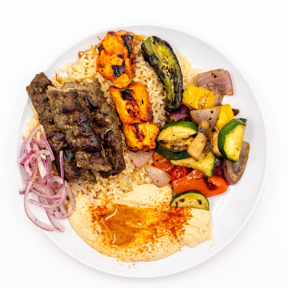 Grilled kebabs, chicken, vegetables, rice, hummus, and sliced red onions arranged on a white plate.