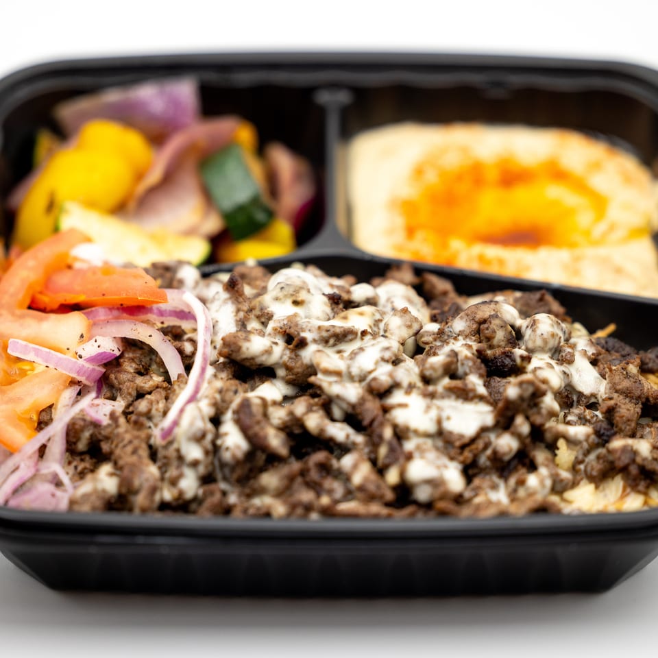 Takeout container filled with seasoned meat topped with creamy sauce, sliced tomatoes and onions, vegetables, and flatbread.