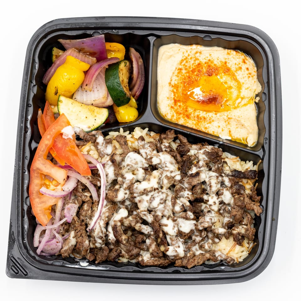 Takeout tray with seasoned rice, grilled meat drizzled with sauce, roasted vegetables, tomatoes, onions, and hummus topped with paprika.