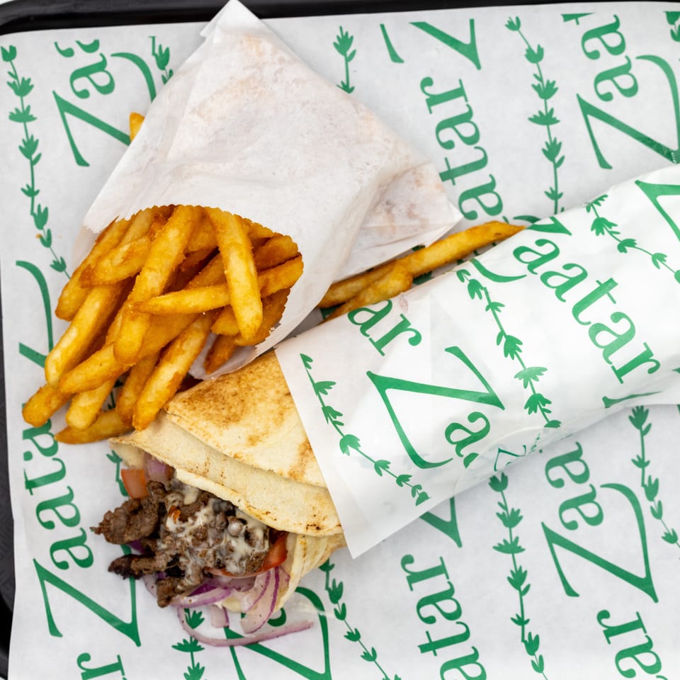 Gyro pita sandwich with grilled meat, onions, tomatoes, and sauce served alongside golden fries on branded paper.