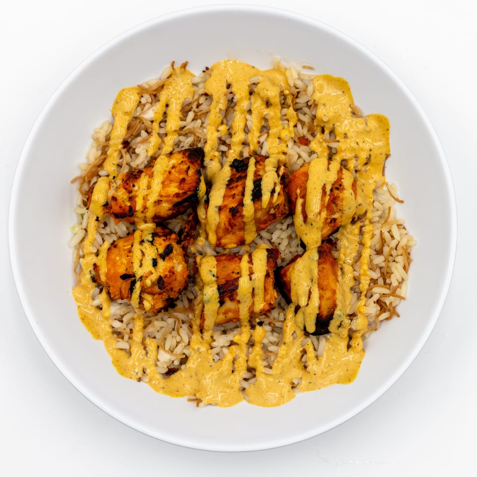 Grilled chicken pieces over seasoned rice, drizzled with creamy yellow sauce on a white plate.