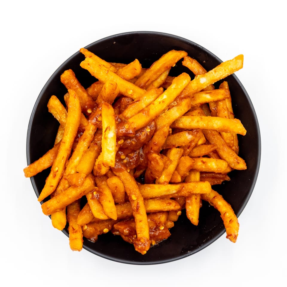 Crispy golden fries coated in spicy sauce and chili flakes fill a black bowl against a white background.