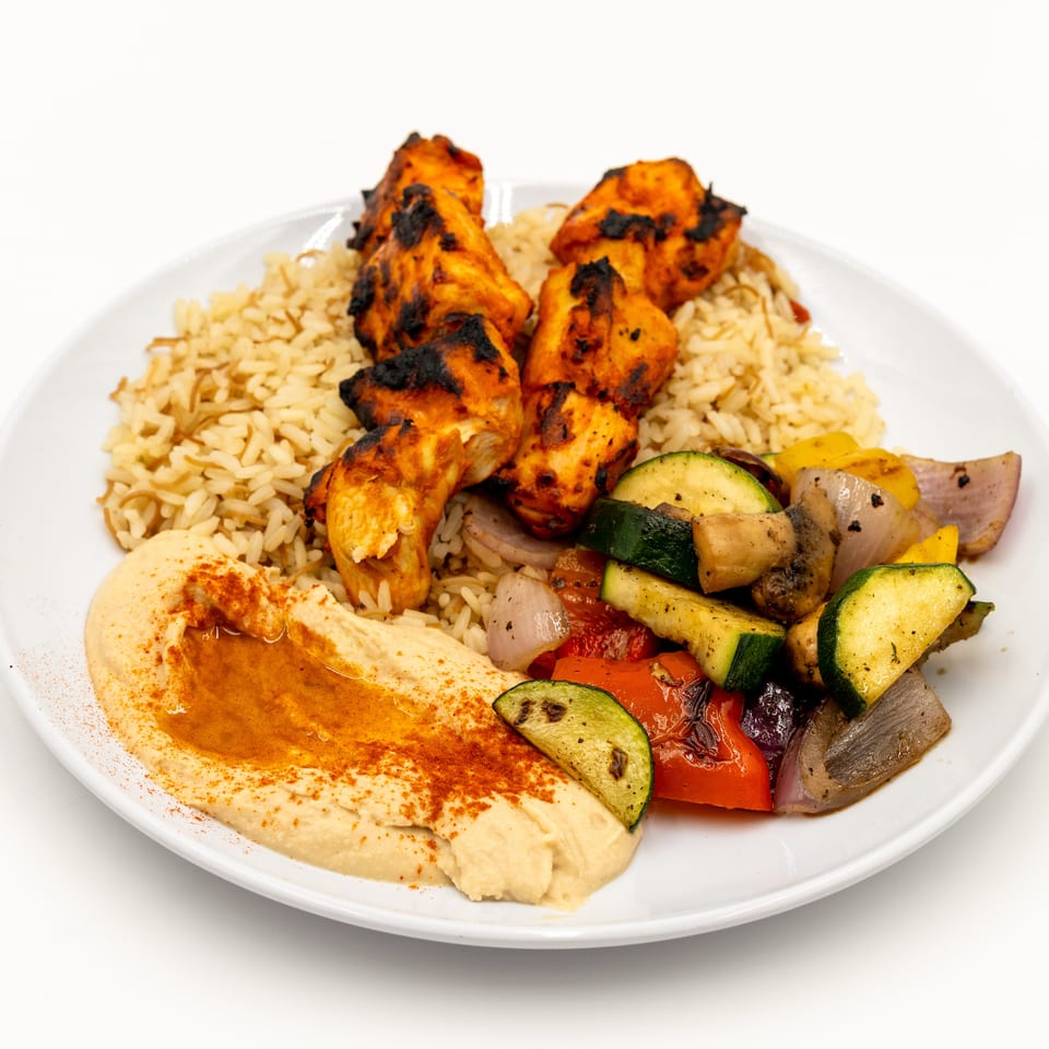 Grilled chicken skewers served with seasoned rice, hummus, and roasted zucchini, peppers, onions, and eggplant on a white plate.
