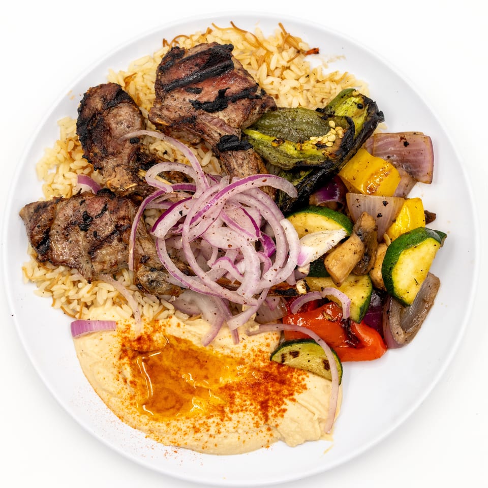 Grilled meat, rice, hummus, roasted vegetables, and sliced red onions arranged on a white plate.